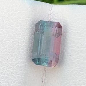 May include: A rectangular gemstone with a gradient of colours, including teal, pink, and clear sections. The gem is faceted with a polished surface, showcasing its internal structure. The stone is set against a white background.