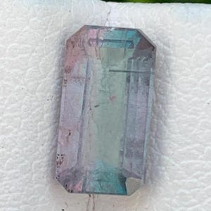 May include: A multi-coloured rectangular gemstone. The gem displays pink, blue, and green hues. It has a faceted cut, with a rectangular shape and beveled edges. The gem is set against a white background.