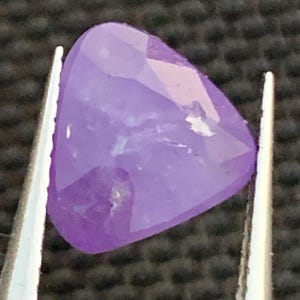 Könnte beinhalten: Ein facettierter, dreieckiger, lavendelfarbener Edelstein wird von einer Pinzette gehalten. Der Edelstein hat eine polierte Oberfläche mit mehreren Facetten, die das Licht reflektieren. Die Farbe ist ein weiches, helles Lila.