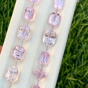 May include: A collection of light pink gemstones in various shapes, including cushion, oval, and pear cuts, are displayed on a white surface. The gemstones are arranged in rows, showcasing their facets and clarity. The background is a blurred green.
