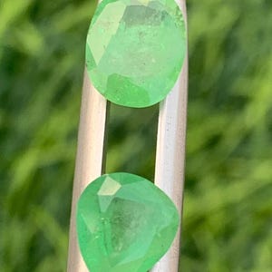 以下が含まれることがあります： 銀色の金属製ピンセットで保持された、鮮やかなライトグリーンのファセットカットルースストーン2個。上の石はオーバルカット、下の石はペアシェイプカットで、どちらも幾何学的なファセットがはっきりと見えます。