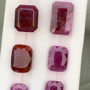 Puede incluir: Una selección de gemas de rubí facetadas en varias formas, incluyendo cortes rectangulares y cojín. Los rubíes muestran una gama de tonos rojos oscuros y rojizo-púrpura, sobre un fondo blanco.