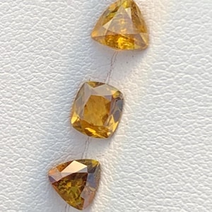 Könnte beinhalten: Drei facettierte Edelsteine in Gelb- und Brauntönen. Zwei sind dreieckig, einer ist kissenförmig geschliffen. Die Edelsteine haben einen Brillantschliff, der das Licht reflektiert. Die Steine liegen auf einer weißen Oberfläche.