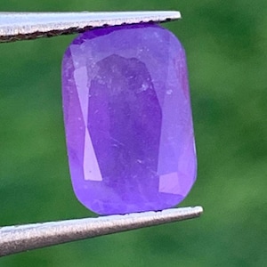 May include: A faceted, rectangular purple gemstone held by silver tweezers. The gem has a polished surface with multiple facets, reflecting light. The background is a blurred green, highlighting the gem's color and shape.