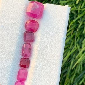 Puede incluir: Una colección de gemas rosas facetadas de varias formas, incluyendo cortes rectangulares, ovalados y redondos. Las gemas se muestran sobre una superficie blanca, con hierba verde visible en el fondo. Las gemas son de un tono rosa vibrante.