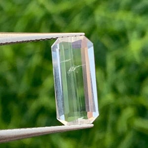 May include: A clear, rectangular gemstone with a faceted cut, held by silver tweezers. The gem has a slight internal flaw and is set against a blurred green background, suggesting natural light and a focus on the gem's clarity.