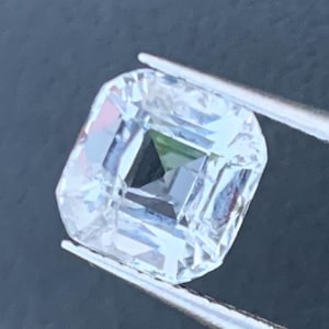 Può includere: Una gemma trasparente, taglio quadrato, tenuta da pinzette argentate. La gemma ha molteplici sfaccettature che riflettono la luce, creando un effetto scintillante. L'interno della gemma ha un'inclusione scura, di forma quadrata. Lo sfondo è grigio scuro.