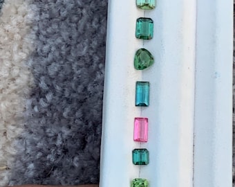 May include: A collection of colorful gemstones, including green, blue, pink, and black tourmalines, arranged on a white surface. The gemstones vary in shape, from rectangular to triangular, and are displayed in a vertical line.