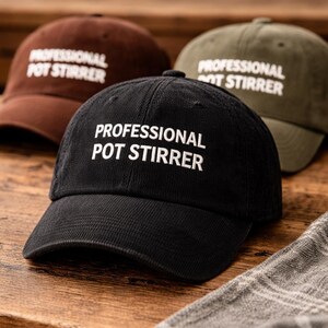 May include: Three corduroy baseball caps in brown, black, and olive green, each embroidered with the text "PROFESSIONAL POT STIRRER" in white. The caps are displayed on a wooden surface with a plaid cloth in the foreground.