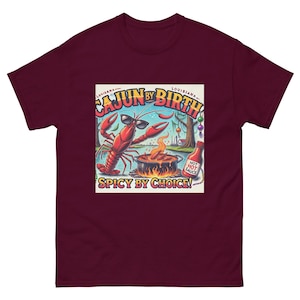 Puede incluir: Una camiseta de color granate con un gráfico de un cangrejo de dibujos animados con gafas de sol y sosteniendo una botella de salsa picante. El texto en el gráfico dice "Cajun by Birth, Spicy by Choice!"