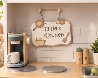 Può includere: Una cucina giocattolo in legno con un cartello appeso che recita Effie's Kitchen, una macchina per il caffè con una tazza bianca, un fornello con tre manopole, un lavello con rubinetto e saliera e pepiera etichettate S e P su piastrelle bianche.