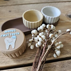 May include: A tan container with the text "JIMMY'S FIRST TOOTH" and a cartoon tooth illustration. Two small, ribbed bowls sit on a brown tray. A sprig of white berries adds a decorative touch.