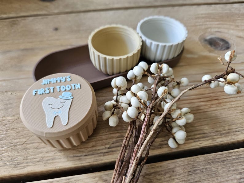May include: A tan container with the text "JIMMY'S FIRST TOOTH" and a cartoon tooth illustration. Two small, ribbed bowls sit on a brown tray. A sprig of white berries adds a decorative touch.