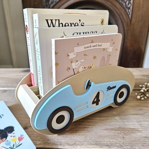 May include: A light blue and beige wooden book organizer shaped like a vintage race car. The car has black wheels, the number 4, and the name "Rome" on the side. Several children's books are inside the organizer.