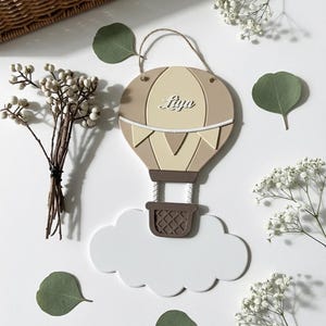 Könnte beinhalten: Wanddekoration in Form eines beigen und cremefarbenen Heißluftballons mit dem Namen "Lilya" in Schreibschrift. Der Ballon schwebt über einer weißen Wolke. Das Bild ist mit Zweigen weißer Blumen und grünen Blättern dekoriert.