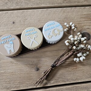 May include: Three small, round keepsake boxes with light blue text and illustrations, reading "Jimmy's First Tooth", "Jimmy's First Curl", and "Jimmy's Umbilical Cord". The boxes are in a dark brown tray, with a sprig of white berries.