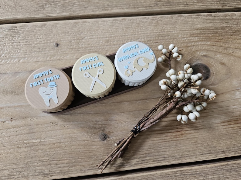 May include: Three small, round keepsake boxes with light blue text and illustrations, reading "Jimmy's First Tooth", "Jimmy's First Curl", and "Jimmy's Umbilical Cord". The boxes are in a dark brown tray, with a sprig of white berries.