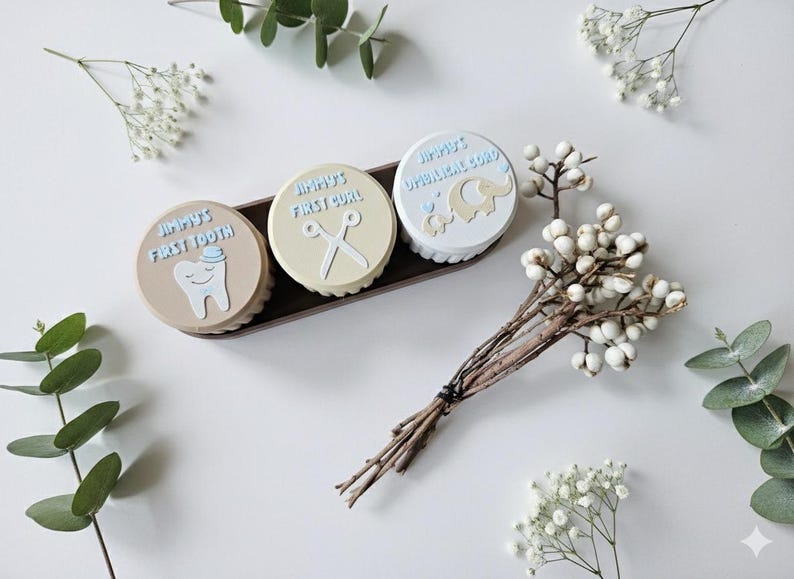 May include: Three round keepsake boxes in a brown tray, each with a light blue border and text. The boxes are labeled "Jimmy's First Tooth", "Jimmy's First Curl", and "Jimmy's Umbilical Cord". Decorative sprigs of white flowers and greenery surround the boxes.