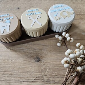May include: Three small, round keepsake boxes with lids, each labeled "Jimmy's First Tooth", "Jimmy's First Curl", and "Jimmy's Umbilical Cord". The boxes are in a brown tray, with a sprig of white berries in the foreground. The lids have light blue text and illustrations.