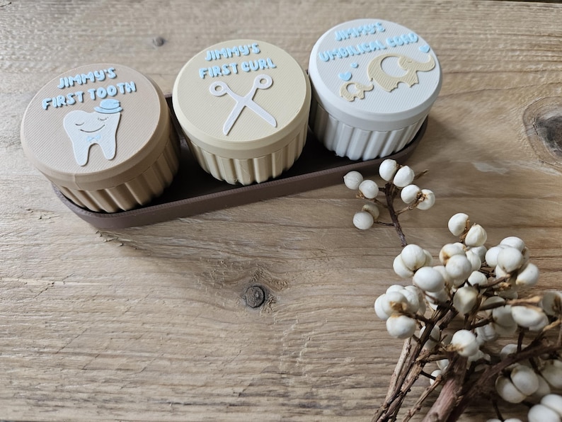 May include: Three small, round keepsake boxes with lids, each labeled "Jimmy's First Tooth", "Jimmy's First Curl", and "Jimmy's Umbilical Cord". The boxes are in a brown tray, with a sprig of white berries in the foreground. The lids have light blue text and illustrations.