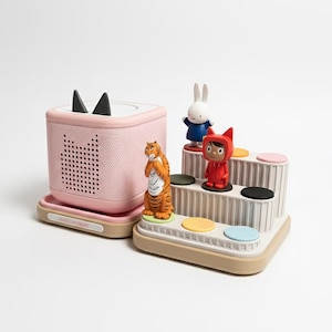 Może przedstawiać: Różowy odtwarzacz audio z czarnymi uszami i kratką głośnika, obok wielopoziomowej zabawki z kolorowymi okręgami i figurkami tygrysa, królika i postaci w czerwonym stroju. Zabawki znajdują się na białej powierzchni.