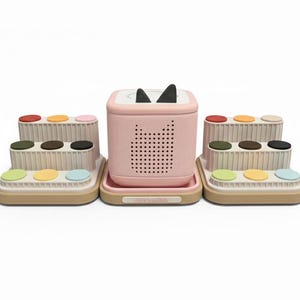 Puede incluir: Un altavoz rosa con orejas de gato y una bandeja, flanqueado por dos soportes beige escalonados con botones de colores. El altavoz tiene una rejilla de agujeros y un dise&ntilde;o de cara de gato. Los soportes tienen botones rojos, amarillos, rosas, verdes, marrones, negros y azules.