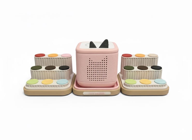 Puede incluir: Un altavoz rosa con orejas de gato y una bandeja, flanqueado por dos soportes beige escalonados con botones de colores. El altavoz tiene una rejilla de agujeros y un dise&ntilde;o de cara de gato. Los soportes tienen botones rojos, amarillos, rosas, verdes, marrones, negros y azules.
