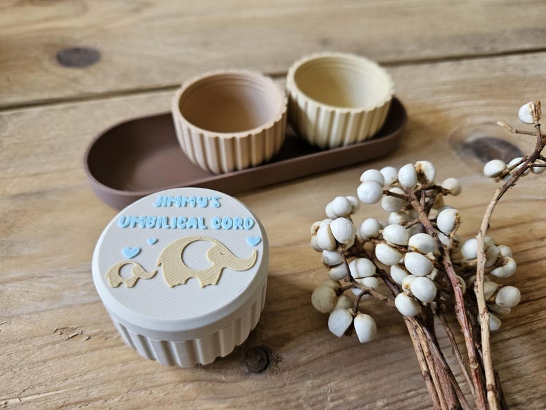 May include: A light blue container with the text "JIMMY'S UMBILICAL CORD" and elephant graphics. Two small beige containers and a brown tray are also visible. A sprig of white berries is on a wooden surface.