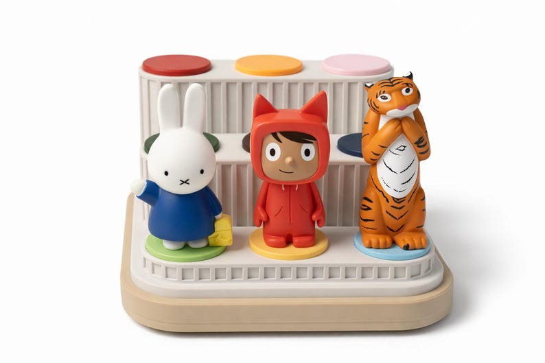 Puede incluir: Un juguete decorativo con tres figuras de dibujos animados: un conejo blanco con un vestido azul, una persona con un mono rojo y un tigre naranja. Las figuras est&aacute;n sobre una plataforma escalonada con acentos circulares de colores. El juguete es beige y blanco.