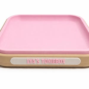 Puede incluir: Una bandeja cuadrada rosa con un borde elevado se encuentra dentro de una base de color beige. La bandeja tiene esquinas redondeadas y una etiqueta blanca con el texto "LYA'S TONIEBOX" en letras rosas.