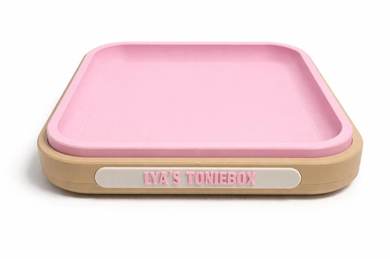 Puede incluir: Una bandeja cuadrada rosa con un borde elevado se encuentra dentro de una base de color beige. La bandeja tiene esquinas redondeadas y una etiqueta blanca con el texto "LYA'S TONIEBOX" en letras rosas.