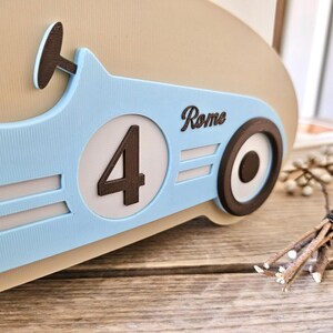 May include: A light blue race car-themed decoration with the number 4 in a white circle. The word "Rome" is written on the side. The car has a brown steering wheel and wheels. The decoration is on a wooden surface.