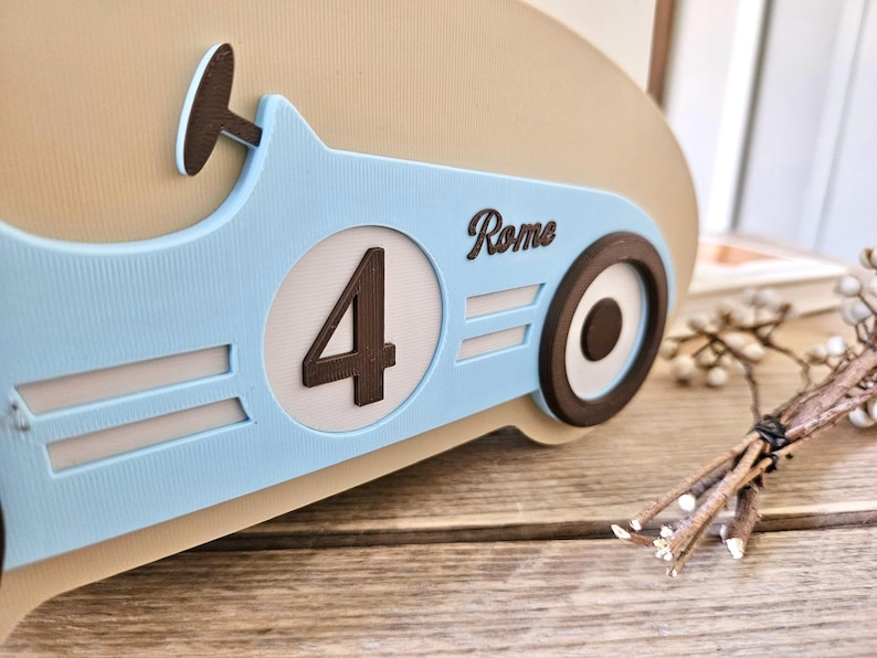 May include: A light blue race car-themed decoration with the number 4 in a white circle. The word "Rome" is written on the side. The car has a brown steering wheel and wheels. The decoration is on a wooden surface.