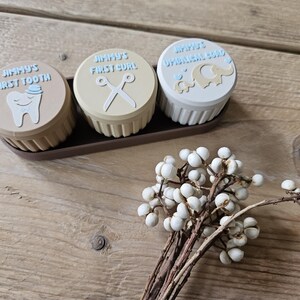 May include: Three small, round keepsake boxes in a brown tray. The lids are beige, with blue text and illustrations: a tooth, scissors, and an elephant. The boxes are labeled "Jimmy's First Tooth", "Jimmy's First Curl", and "Jimmy's Umbilical Cord".