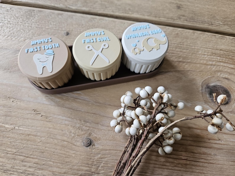 May include: Three small, round keepsake boxes in a brown tray. The lids are beige, with blue text and illustrations: a tooth, scissors, and an elephant. The boxes are labeled "Jimmy's First Tooth", "Jimmy's First Curl", and "Jimmy's Umbilical Cord".