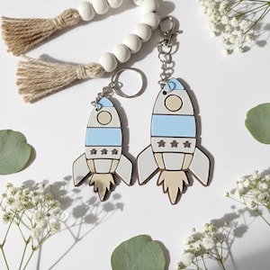 May include: Two rocket-shaped keyrings in light blue and beige. Each keyring has a silver ring and clasp. The rockets feature three stars and a circular window. The background includes white beads, greenery, and white flowers.