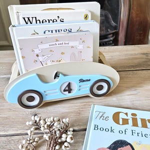 May include: A light blue wooden car-shaped book holder with the number 4 and the name "Rome" on the side. The holder contains several children's books, including one labeled "Where's" and another "touch and feel". A small floral arrangement sits in front.