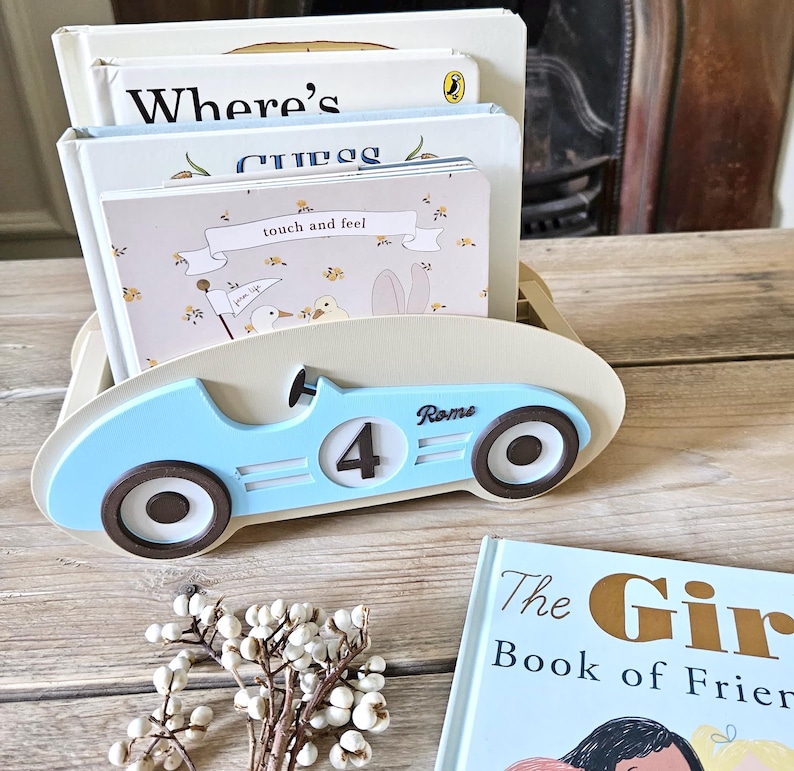 May include: A light blue wooden car-shaped book holder with the number 4 and the name "Rome" on the side. The holder contains several children's books, including one labeled "Where's" and another "touch and feel". A small floral arrangement sits in front.