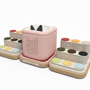 Puede incluir: Un altavoz rosa con orejas de gato y una base a juego, rodeado de varios conjuntos de botones multicolores. El altavoz tiene un dise&ntilde;o perforado y orejas de gato negras. Los conjuntos de botones tienen tres niveles con botones coloridos.