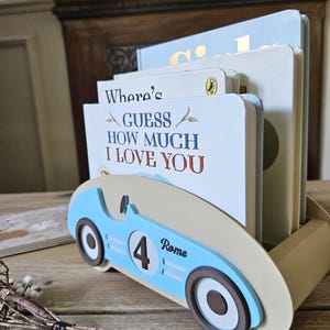 May include: A light blue and tan book holder shaped like a race car, holding several children's books. The book holder has a number 4 and the name "Rome" on the side. The book titles include "Where's Guess How Much I Love You."