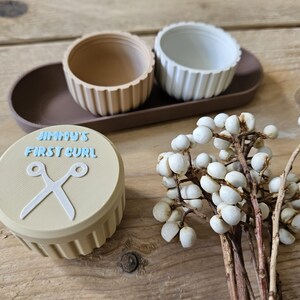 May include: A beige container with the text "JIMMY'S FIRST CURL" and a pair of scissors. Two small, ribbed bowls sit on a brown tray. A sprig of white berries is also present. The items are arranged on a wooden surface.