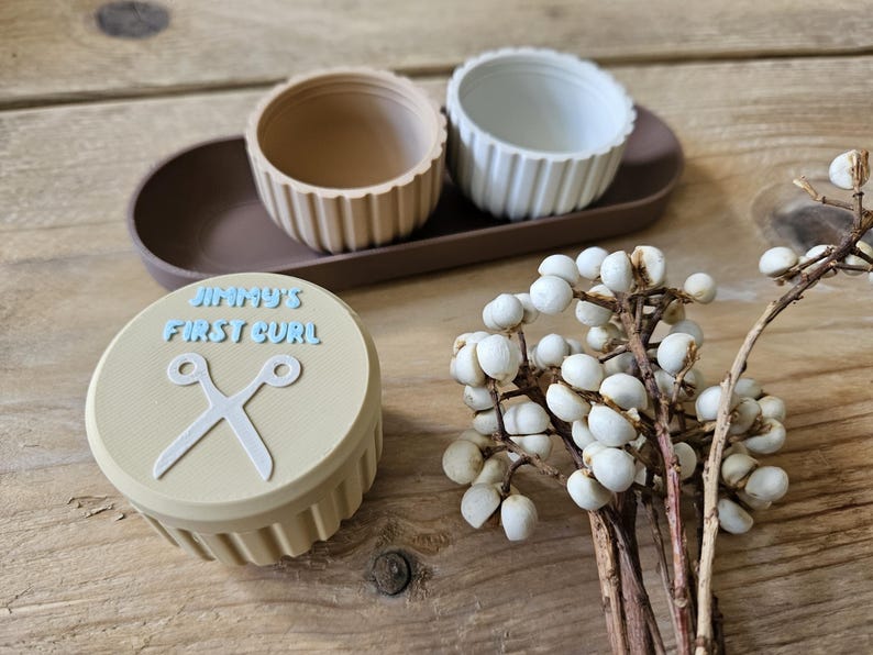 May include: A beige container with the text "JIMMY'S FIRST CURL" and a pair of scissors. Two small, ribbed bowls sit on a brown tray. A sprig of white berries is also present. The items are arranged on a wooden surface.