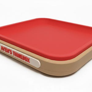 Puede incluir: Una caja de almacenamiento rectangular con interior rojo y exterior beige. La caja tiene esquinas redondeadas y una etiqueta blanca con el texto "AYEN'S TONIEBOX" en rojo. La caja est&aacute; dise&ntilde;ada para guardar objetos.