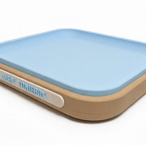 Puede incluir: Un contenedor rectangular con interior azul claro y exterior beige. El contenedor tiene bordes redondeados y una etiqueta blanca con el texto "ALEX TONIEBOX" en azul claro.