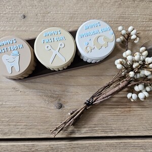 May include: Three round keepsake boxes in a dark brown tray. The boxes are labeled "Jimmy's First Tooth", "Jimmy's First Curl", and "Jimmy's Umbilical Cord". Each box has a different design, including a tooth, scissors, and an elephant. A small floral arrangement is in the foreground.