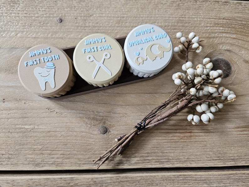May include: Three round keepsake boxes in a dark brown tray. The boxes are labeled "Jimmy's First Tooth", "Jimmy's First Curl", and "Jimmy's Umbilical Cord". Each box has a different design, including a tooth, scissors, and an elephant. A small floral arrangement is in the foreground.