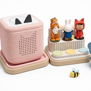 Puede incluir: Un reproductor de audio rosa con orejas de gato negras, un altavoz y una bandeja. Varias figuras de juguete coloridas est&aacute;n dispuestas cerca, incluyendo un tigre, un conejo y una figura con capucha roja. Tambi&eacute;n hay un elefante azul, un dinosaurio verde y una abeja.