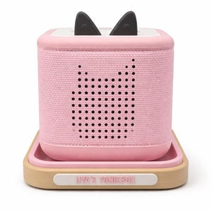 Puede incluir: Un reproductor de audio rosa con orejas de gato negras en la parte superior, sobre una bandeja. El reproductor tiene un dise&ntilde;o de cara de gato hecho de agujeros. La bandeja es beige con un borde rosa y el texto "LIA'S TONIEBOX" en la parte delantera.