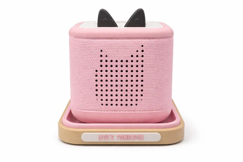 Puede incluir: Un reproductor de audio rosa con orejas de gato negras en la parte superior, sobre una bandeja. El reproductor tiene un dise&ntilde;o de cara de gato hecho de agujeros. La bandeja es beige con un borde rosa y el texto "LIA'S TONIEBOX" en la parte delantera.