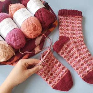 Pode incluir: Meias de malha com um padrão geométrico em tons de rosa, vermelho e pêssego. Vários novelos de lã em cores correspondentes são visíveis, juntamente com uma meia parcialmente tricotada e agulhas de tricô. O fio é rotulado como "Scheepjes Metropolis".